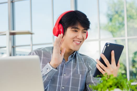 Asian young business man happiness with red headphones he sitting on desk workplace using digital tablet while greeting by video chat conferences talking and discussing