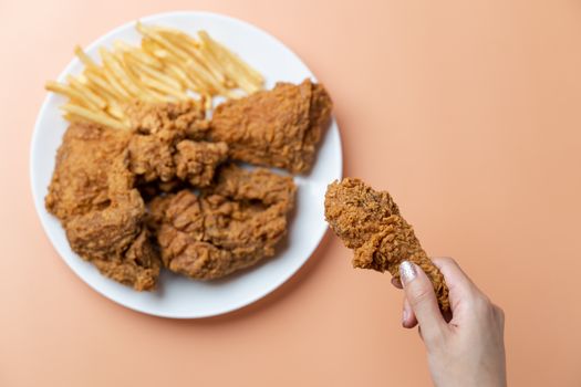 Hand holding drumsticks, crispy fried chicken with french fries in white plate on orange background.