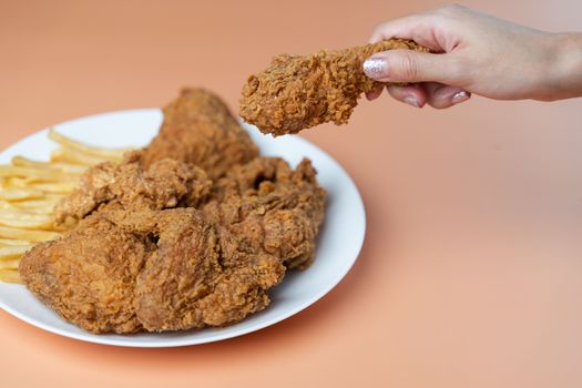 Hand holding drumsticks, crispy fried chicken with french fries in white plate on orange background.