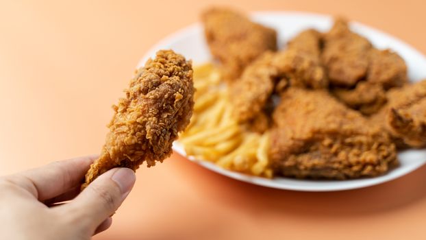 Hand holding drumsticks, crispy fried chicken with french fries in white plate on orange background.