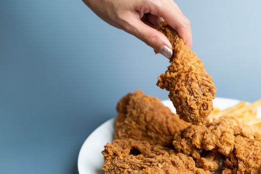 Hand holding drumsticks, crispy fried chicken in white plate on blue background.