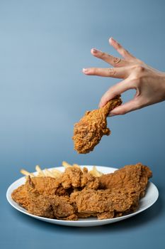 Hand holding drumsticks, crispy fried chicken in white plate on blue background.