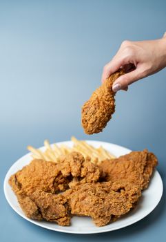 Hand holding drumsticks, crispy fried chicken with french fries in white plate on blue background.