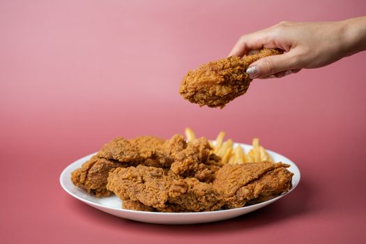 Hand holding drumsticks, crispy fried chicken with french fries in white plate on red background.