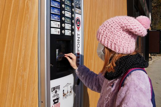 Varna, Bulgaria - November, 26, 2020: teen girl in a medical mask buys a drink from a vending machine on the street