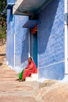 Jodhpur, India - December 9, 2019: An Indian woman in colorful clothes sitting in front of a blue house.