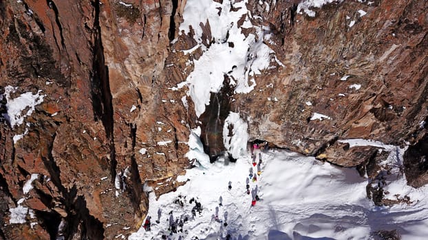 Freezing waterfall in the snowy mountains. View from the drone, from above. The rocks are covered with snow and ice. A small stream of water runs. The waterfall freezes. A group of people are resting