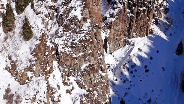 Freezing waterfall in the snowy mountains. View from the drone, from above. The rocks are covered with snow and ice. A small stream of water runs. The waterfall freezes. A group of people are resting