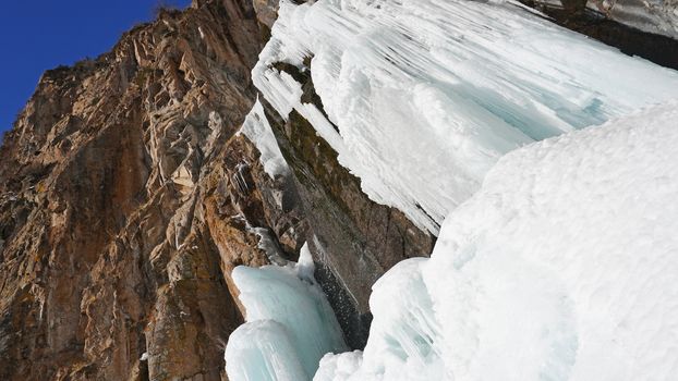Frozen waterfall among the rocks. The waterfall is freezing, huge icicles. Ice white and blue. Brown rocks and splashes of water. Winter waterfall. White snow and blue sky. Water runs down the ice.
