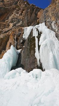 Frozen waterfall among the rocks. The waterfall is freezing, huge icicles. Ice white and blue. Brown rocks and splashes of water. Winter waterfall. White snow and blue sky. Water runs down the ice.