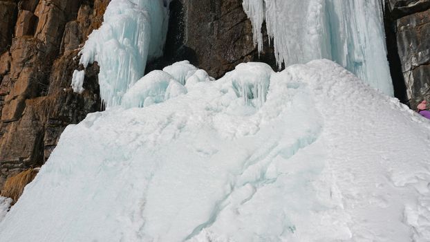 Frozen waterfall among the rocks. The waterfall is freezing, huge icicles. Ice white and blue. Brown rocks and splashes of water. Winter waterfall. White snow and blue sky. Water runs down the ice.