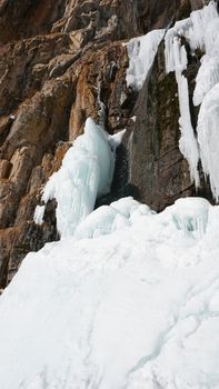 Frozen waterfall among the rocks. The waterfall is freezing, huge icicles. Ice white and blue. Brown rocks and splashes of water. Winter waterfall. White snow and blue sky. Water runs down the ice.