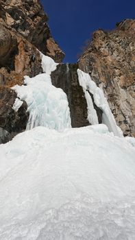 Frozen waterfall among the rocks. The waterfall is freezing, huge icicles. Ice white and blue. Brown rocks and splashes of water. Winter waterfall. White snow and blue sky. Water runs down the ice.