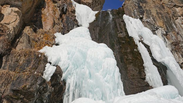 Frozen waterfall among the rocks. The waterfall is freezing, huge icicles. Ice white and blue. Brown rocks and splashes of water. Winter waterfall. White snow and blue sky. Water runs down the ice.