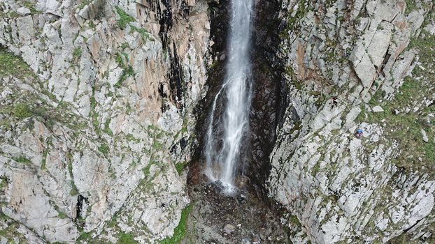 High waterfall among the rocks. View from a drone. Large rocks and steep slopes surround the mountain waterfall. Trees and grass grow below. Clouds float by. The view from top. Ala Archa, Kyrgyzstan.