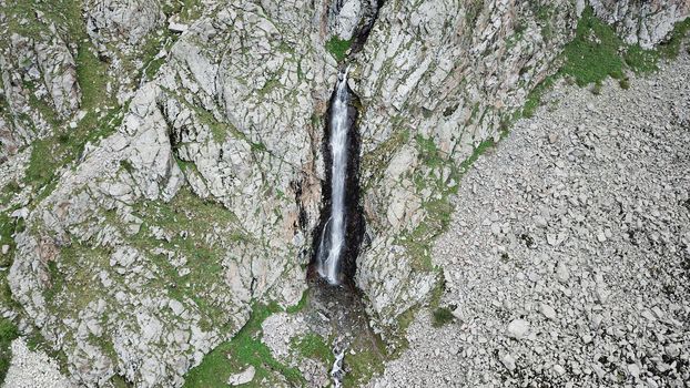 High waterfall among the rocks. View from a drone. Large rocks and steep slopes surround the mountain waterfall. Trees and grass grow below. Clouds float by. The view from top. Ala Archa, Kyrgyzstan.