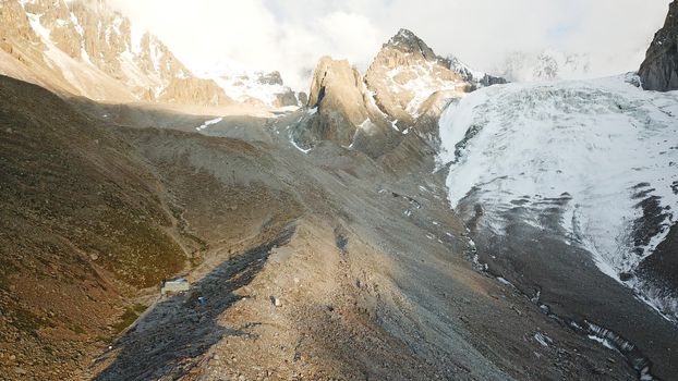 Glacier covered with rocks. Great cloud. Mountainous terrain, lots of big rocks, high mountains and snowy peaks. Huge ice covered the gorge. Small house is visible. Ratsek hut at an altitude of 3200m.
