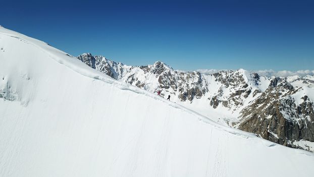 A group of climbers climb to the top of the peak. Dangerous, steep slope. Avalanche-prone area. Mountains covered with snow. Steep cliffs and large rocks. Above the mountains, top view from a drone.