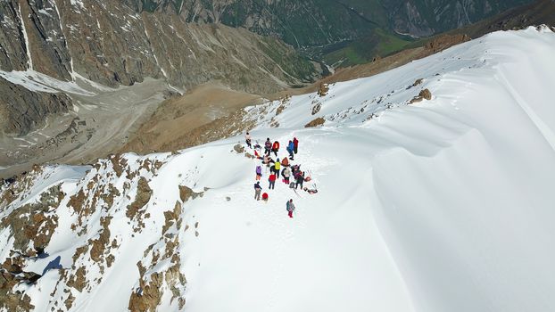 A group of climbers climb to the top of the peak. Dangerous, steep slope. Avalanche-prone area. Mountains covered with snow. Steep cliffs and large rocks. Above the mountains, top view from a drone.
