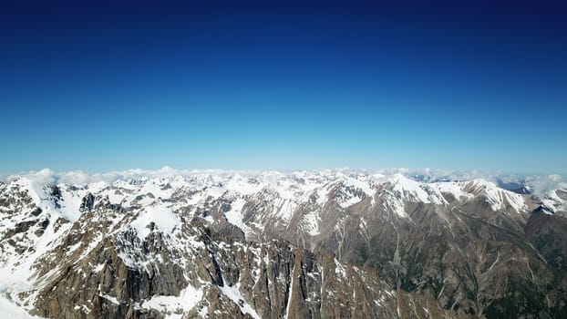 Snow-capped mountains and blue sky. View from a drone. Huge peaks covered with snow, steep cliffs. Blue, clear sky above the mountains. Flying over the peaks. Below you can see green trees and tarva.