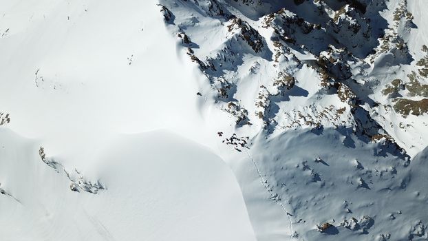 A group of climbers climb to the top of the peak. Dangerous, steep slope. Avalanche-prone area. Mountains covered with snow. Steep cliffs and large rocks. Above the mountains, top view from a drone.