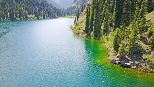Kolsay lake among green hills and mountains. The mountain lake is surrounded by green forest, tall coniferous trees, grass and bushes. Clean water is like a mirror. Tourists swim on boats. Kazakhstan