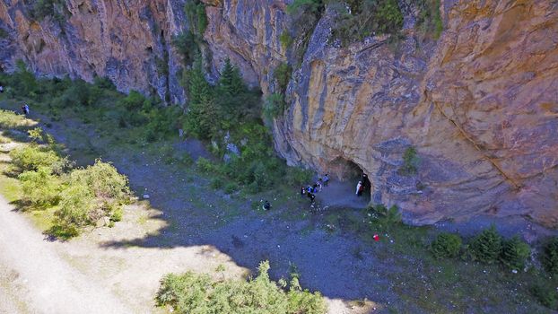 Top view of the cave, forest, rocks and road. A group of tourists walking along the trail. There is a huge cave in the rock. Green coniferous forest, trees and grass grow on the rocks. The river runs.