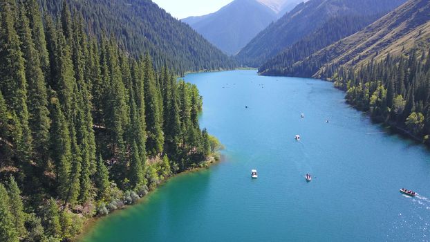 Kolsay lake among green hills and mountains. The mountain lake is surrounded by green forest, tall coniferous trees, grass and bushes. Clean water is like a mirror. Tourists swim on boats. Kazakhstan