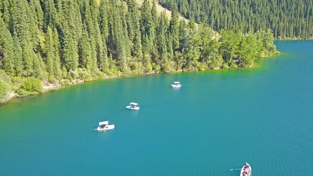 Kolsay lake among green hills and mountains. The mountain lake is surrounded by green forest, tall coniferous trees, grass and bushes. Clean water is like a mirror. Tourists swim on boats. Kazakhstan