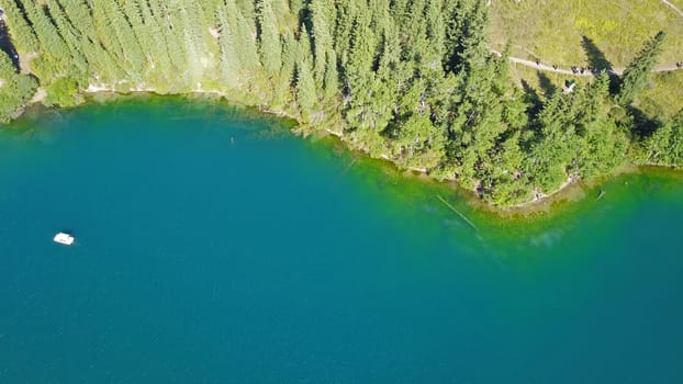 Kolsay lake among green hills and mountains. The mountain lake is surrounded by green forest, tall coniferous trees, grass and bushes. Clean water is like a mirror. Tourists swim on boats. Kazakhstan