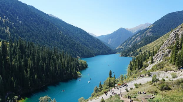 Kolsay lake among green hills and mountains. The mountain lake is surrounded by green forest, tall coniferous trees, grass and bushes. Clean water is like a mirror. Tourists swim on boats. Kazakhstan