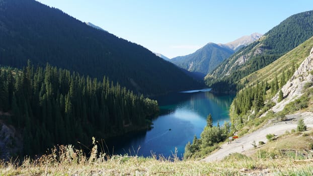 Kolsay lake among green hills and mountains. The mountain lake is surrounded by green forest, tall coniferous trees, grass and bushes. Clean water is like a mirror. Tourists swim on boats. Kazakhstan