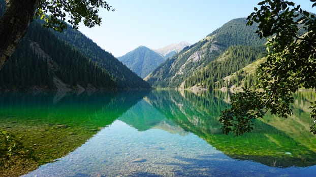 Kolsay lake among green hills and mountains. The mountain lake is surrounded by green forest, tall coniferous trees, grass and bushes. Clean water is like a mirror. Tourists swim on boats. Kazakhstan