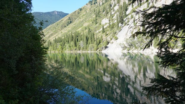 Kolsay lake among green hills and mountains. The mountain lake is surrounded by green forest, tall coniferous trees, grass and bushes. Clean water is like a mirror. Tourists swim on boats. Kazakhstan