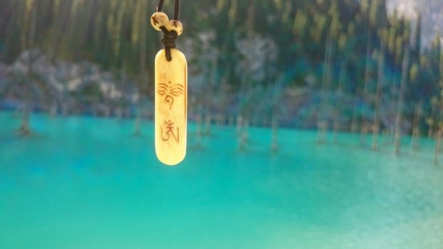 Macro symbol OM (AUM). A locket with OM symbol. The background is blurred. Coniferous trees, a lake, fir branches, blue and green water of the lake are visible. Mountain lake. Kazakhstan, Almaty.