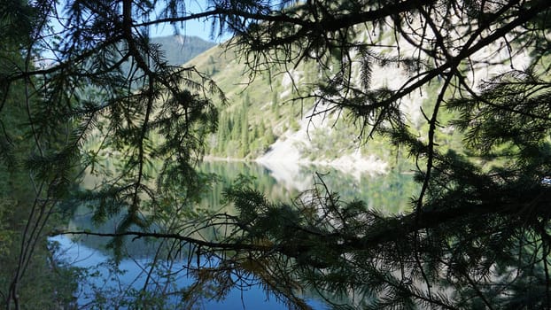 Kolsay lake among green hills and mountains. The mountain lake is surrounded by green forest, tall coniferous trees, grass and bushes. Clean water is like a mirror. Tourists swim on boats. Kazakhstan