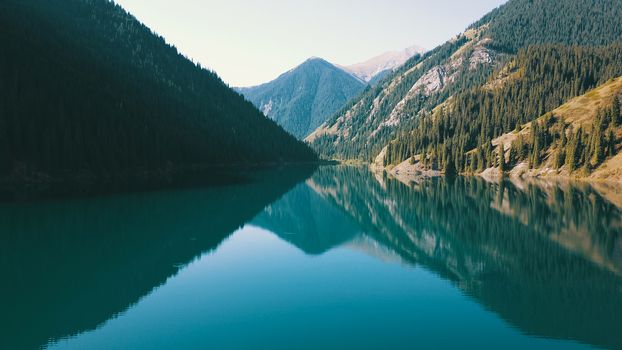 Kolsay lake among green hills and mountains. The mountain lake is surrounded by green forest, tall coniferous trees, grass and bushes. Clean water is like a mirror. Tourists swim on boats. Kazakhstan