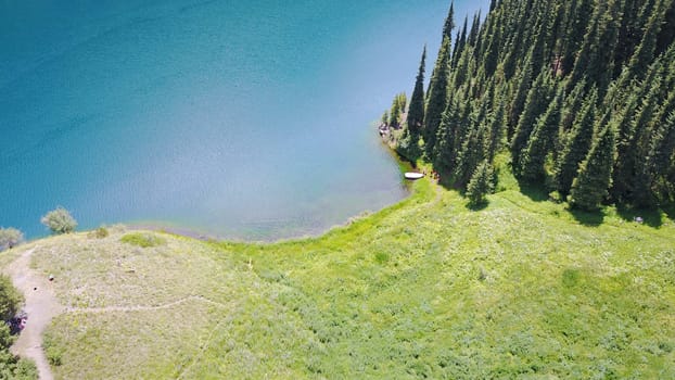 Kolsay lake among green hills and mountains. The mountain lake is surrounded by green forest, tall coniferous trees, grass and bushes. Clean water is like a mirror. Tourists swim on boats. Kazakhstan