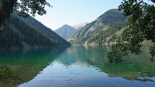 Kolsay lake among green hills and mountains. The mountain lake is surrounded by green forest, tall coniferous trees, grass and bushes. Clean water is like a mirror. Tourists swim on boats. Kazakhstan