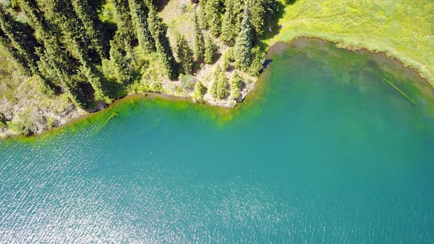 Kolsay lake among green hills and mountains. The mountain lake is surrounded by green forest, tall coniferous trees, grass and bushes. Clean water is like a mirror. Tourists swim on boats. Kazakhstan