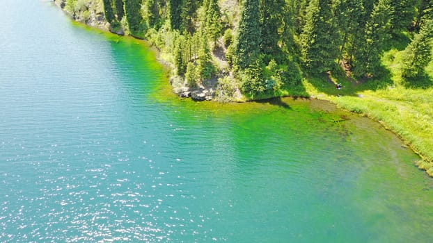 Kolsay lake among green hills and mountains. The mountain lake is surrounded by green forest, tall coniferous trees, grass and bushes. Clean water is like a mirror. Tourists swim on boats. Kazakhstan