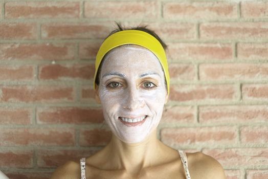 Relaxed woman applies mineral clay mask on face for rejuvenation, has headband on head