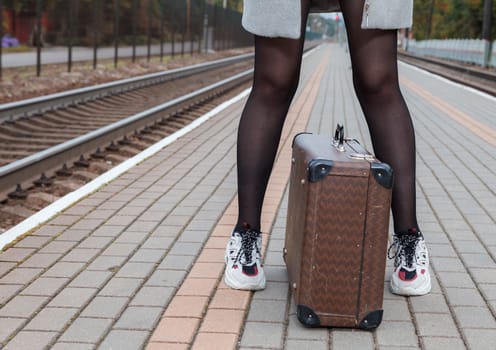 woman in a gray coat and with a suitcase standing at the railway station on an autumn day. legs closeup