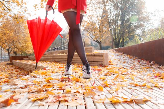 woman in red coat and umbrella standing on alley of city park on sunny autumn day. legs closeup