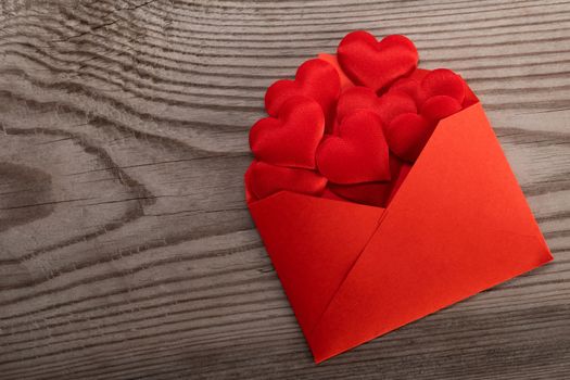 Valentine day love letter, envelope of craft red paper with red hearts heap spread on wood background.