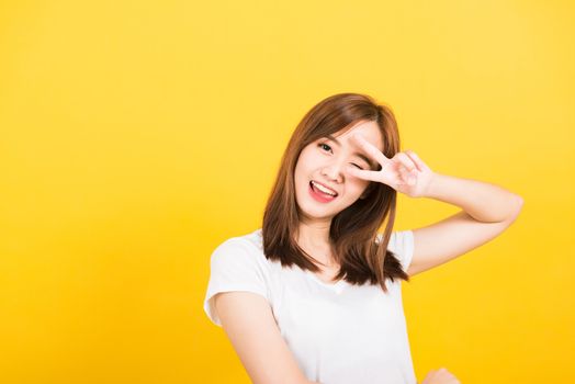 Asian happy portrait beautiful cute young woman teen smile standing wear t-shirt showing finger making v-sign symbol near eye  looking to camera isolated, studio shot yellow background with copy space