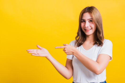 Asian Thai happy portrait beautiful young woman standing to hold something on palm away side and point the finger to it looking to camera, studio shot isolated on yellow background with copy space