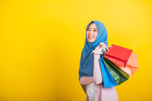 Asian Muslim Arab, Portrait of happy beautiful young woman Islam religious wear veil hijab funny smile she holding colorful shopping bags, studio shot isolated on yellow background