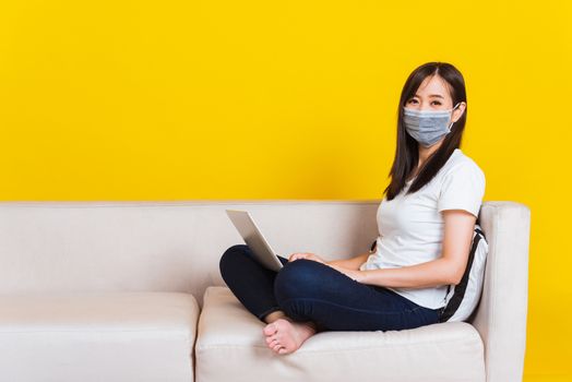 Portrait Asian of beautiful young woman sitting on sofa wearing medical face mask protective she work from home with laptop computer during Coronavirus studio shot isolated on yellow background