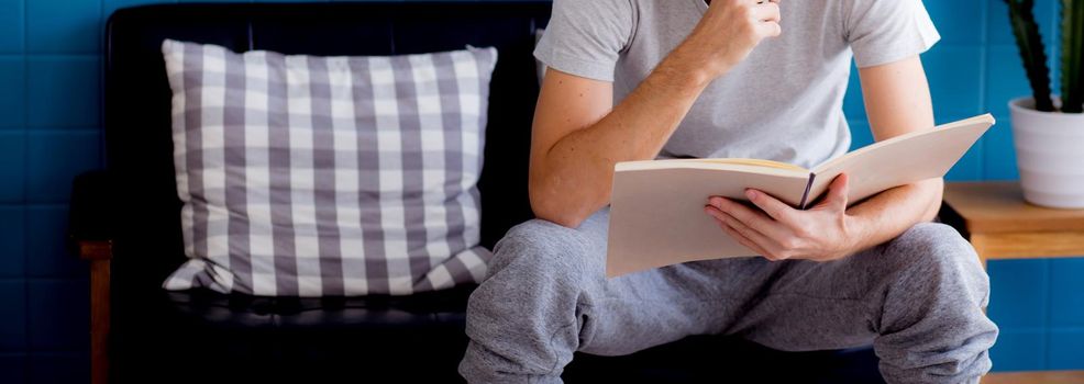 Young man reading a book sitting on sofa for rest is hobby in living room, stay home for leisure, lifestyle of new normal for social distracting, male learn and study in interesting in spare time.